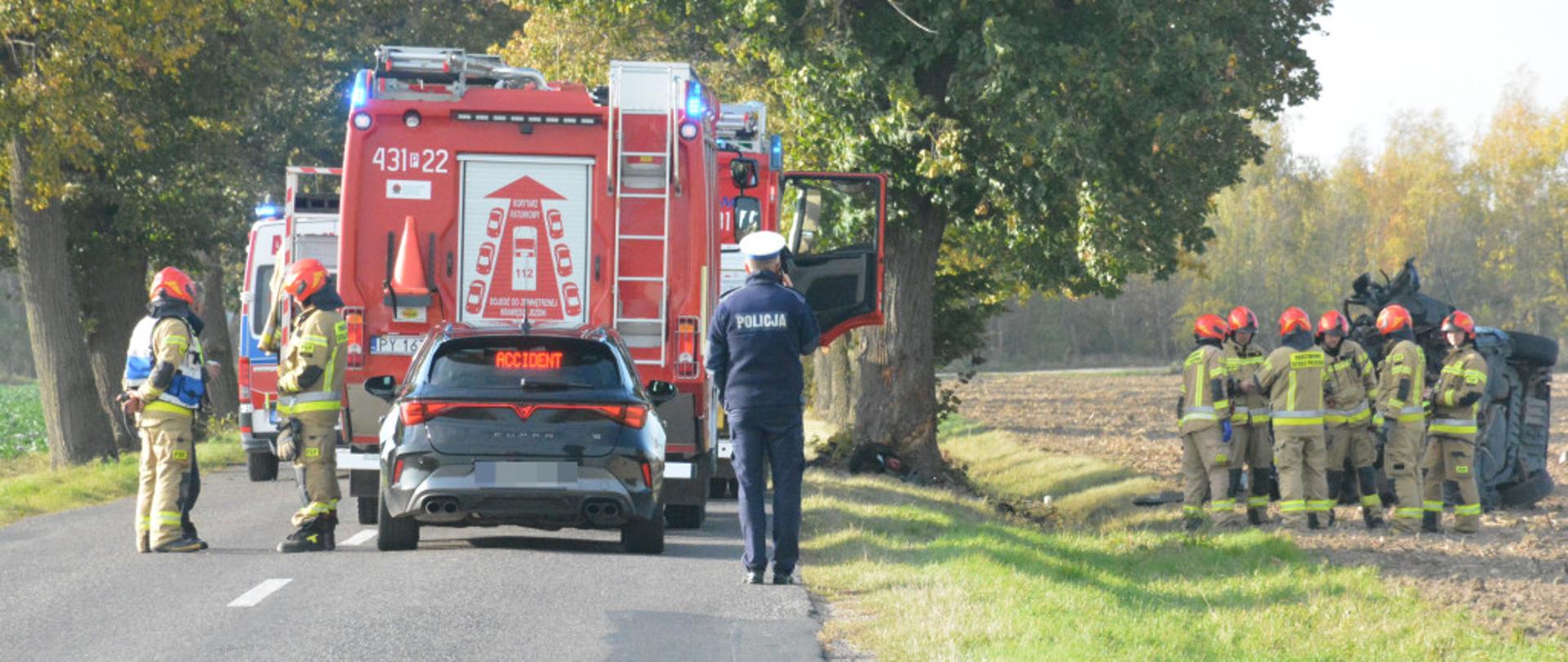 Na zdjęciu znajdują się policjanci oraz strażacy. Po prawej stronie leży uszkodzony pojazd. Po lewej stronie stoi radiowóz oraz pojazdy pożarnicze.