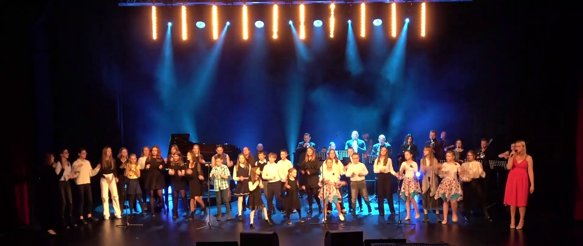 Zdjęcie przedstawiające chór szkolny prowadzony przez mgr Celestynę Koszak oraz zespół Combo prowadzony przez mgr Bogusława Kłaka w trakcie finałowej piosenki podczas Koncertu Jubileuszowego z okazji 40-lecia powstania szkoły. Koncert odbył się w sali kina "Forum" BOK-MCC w dniu 14 grudnia 2023 r. 