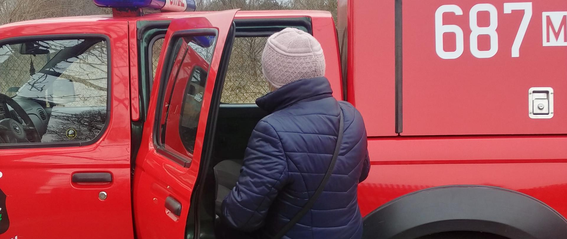 Na pierwszym planie na zdjęciu widać samochód OSP Ślężany typu pic-up. Do środka wsiada kobieta, która ma założoną maseczkę na twarzy. 