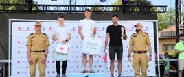 Na zdjęciu widać ceremonię wręczenia nagród. Trzech sportowców stoi na podium, otrzymując trofea i upominki. Dwóch mężczyzn w mundurach stoi po bokach podium.