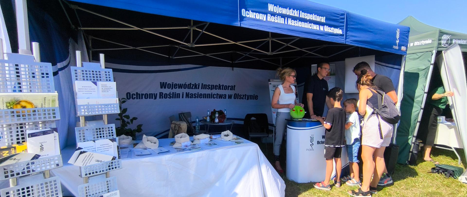 Stoisko targowe, duży namiot, po prawej: za stolikiem stoją dwie osoby a naprzeciw nich grupa kilku osób, po lewej: stolik z rozłożonymi ulotkami