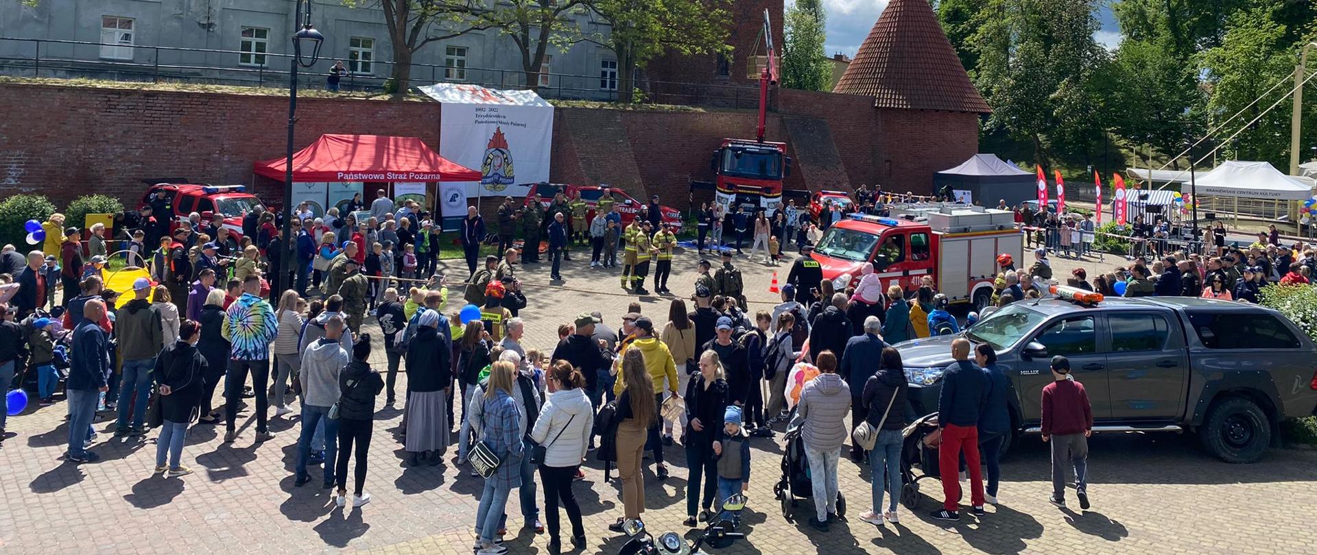 Zdjęcie zrobione z góry. Tłum ludzi zgromadzony na placu. W okół rozstawiony sprzęt i samochody. W oddali budynek. Z prawej u góry niebieskie niebo, reszta białe chmury.