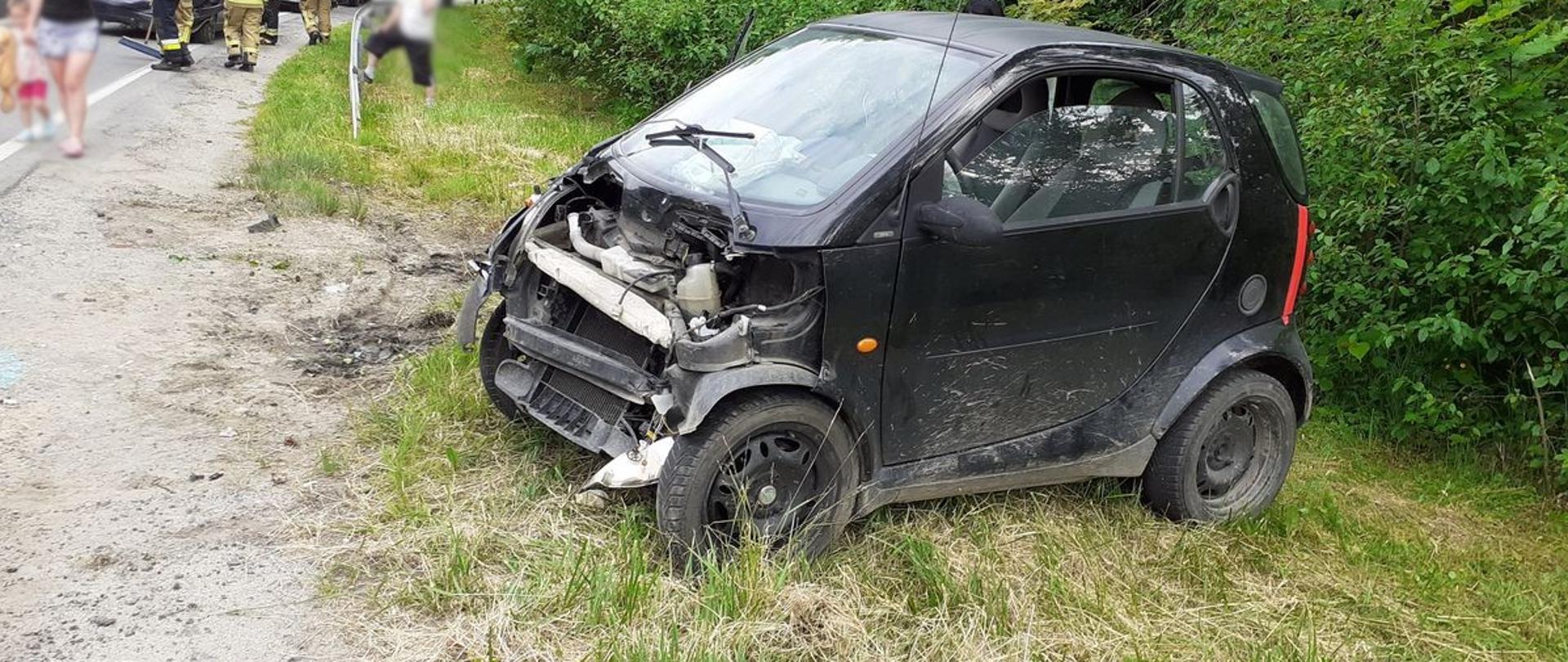 Samochód z uszkodzonym przodem na poboczu. Na drodze ludzie, strażacy oraz samochody służb ratowniczych.