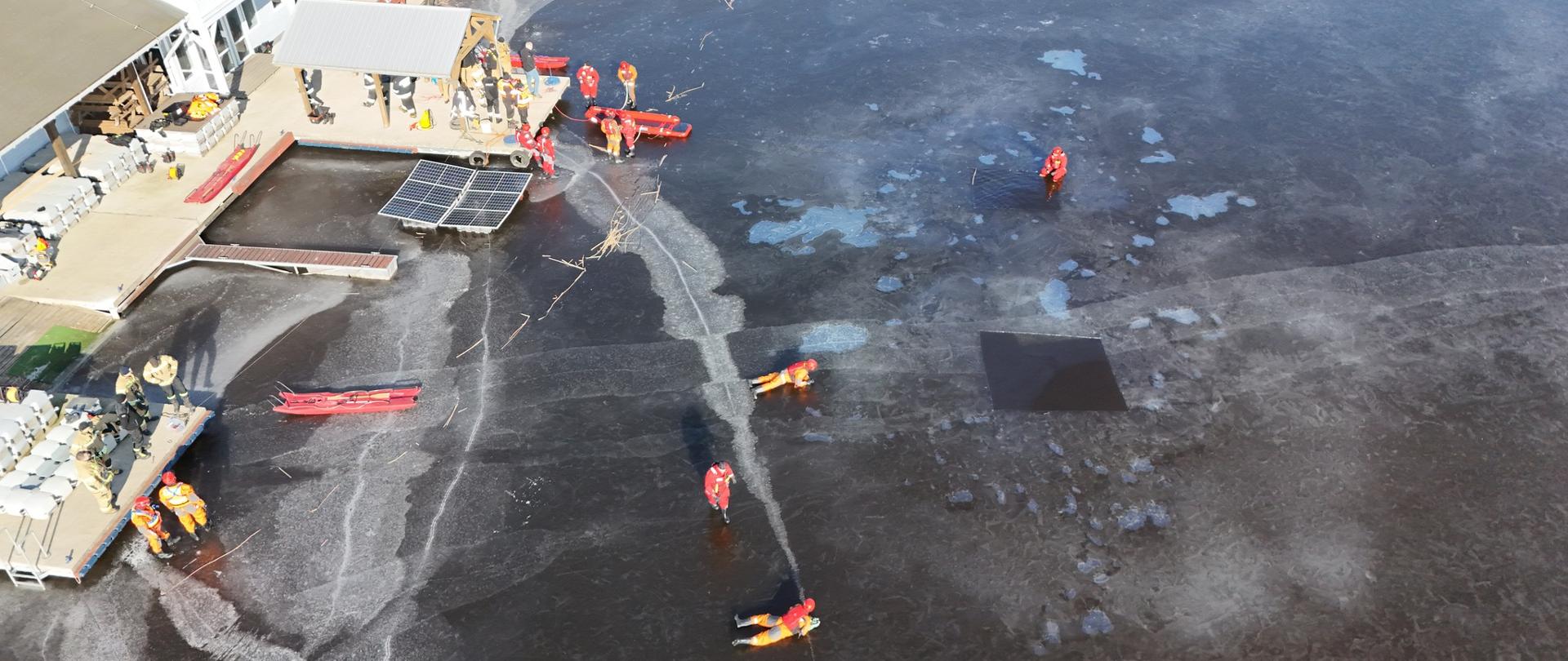 Zdjęcie przedstawia strażaków ćwiczących ratownictwo lodowe na jeziorze.
W tle jezioro o las.
