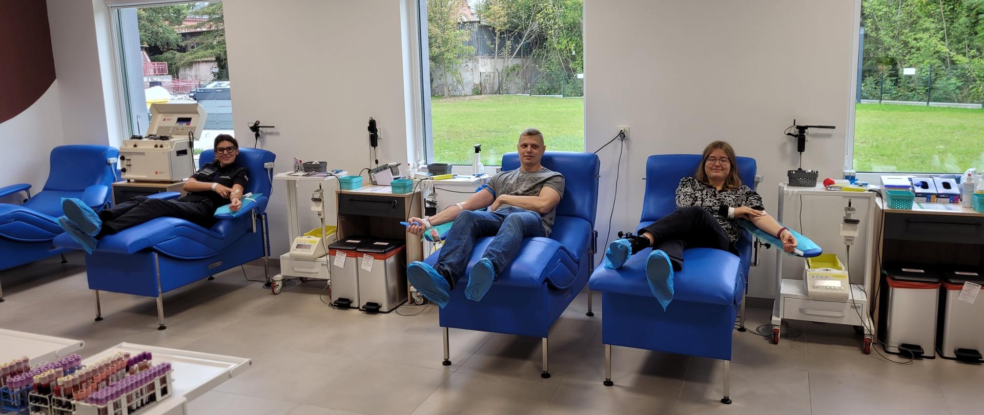 Jasne pomieszczenie, w którym ustawione są niebieskie fotele. Dwie kobiety i jeden mężczyzna leżą na nich w trakcie oddawania krwi. W tle tereny zielone widziane przez okna.