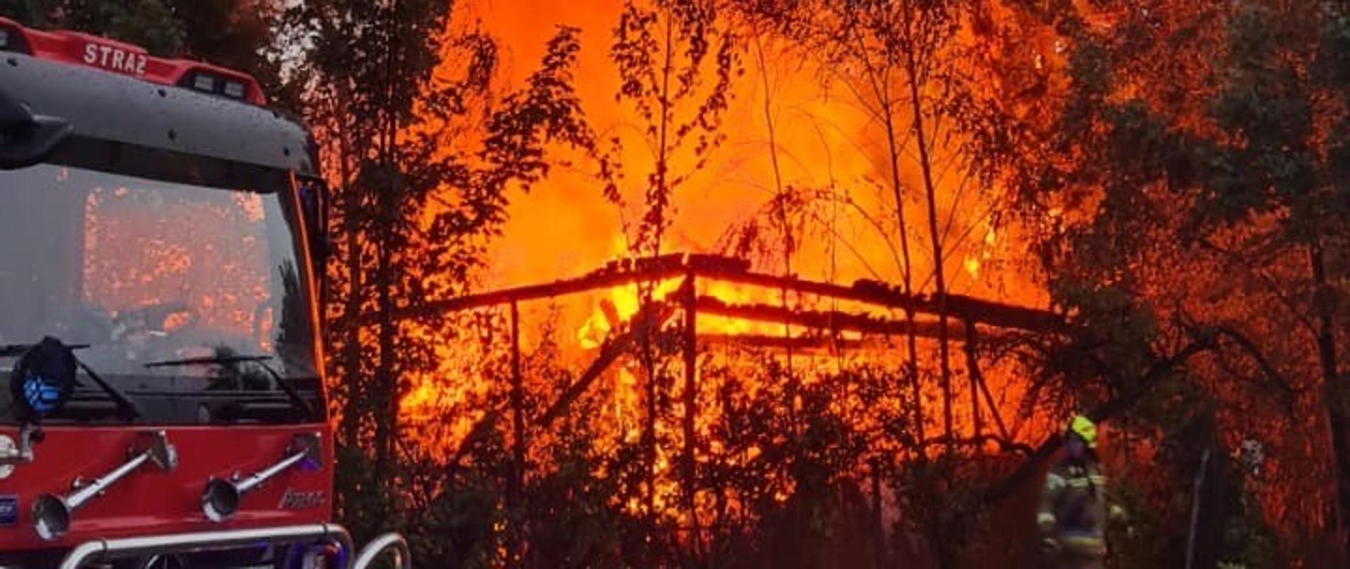 Strażacy w trakcie akcji gaśniczej rozwiniętego pożaru budynku gospodarczego. Na pierwszym planie wóz strażacki, w tle wysoka kolumna gęstego, czarnego dymu i intensywne płomienie ogarniające drewnianą konstrukcję budynku, otoczoną przez drzewa