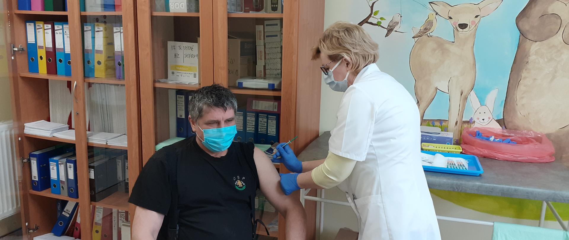 Widać strażaka siedzącego na krześle. Pielęgniarka aplikuje strażakowi szczepionkę w lewe ramię. W tle widać gablotę z segregatorami.