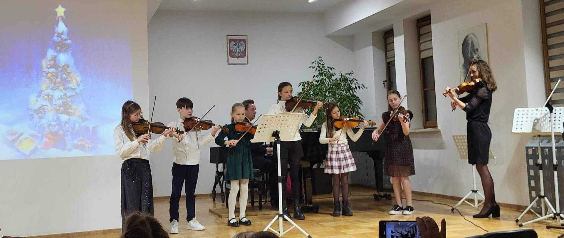 na estradzie grają uczniowie klasy skrzypiec, z lewej projektor z wyświetlanym plakatem ubranej choinki w zimowej aurze, po prawej nauczyciel gra na skrzypcach oraz w tylnej części nauczyciel towarzyszy w grze na fortepianie