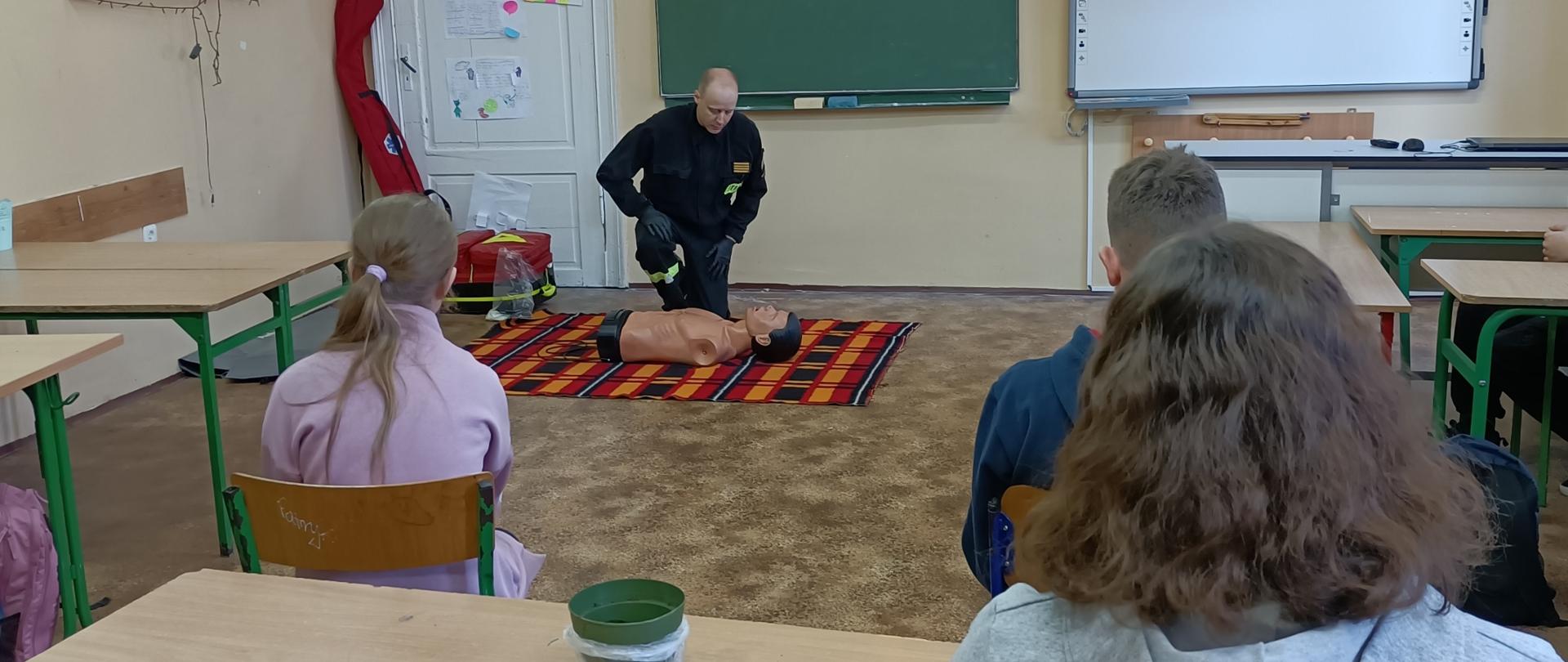 Strażak pochylony nad fantomem tłumaczy dla uczniów siedzących w ławkach zasady udzielania KPP. Za strażakiem widoczne są dwie tablice do pisania oraz wyjście z sali. 