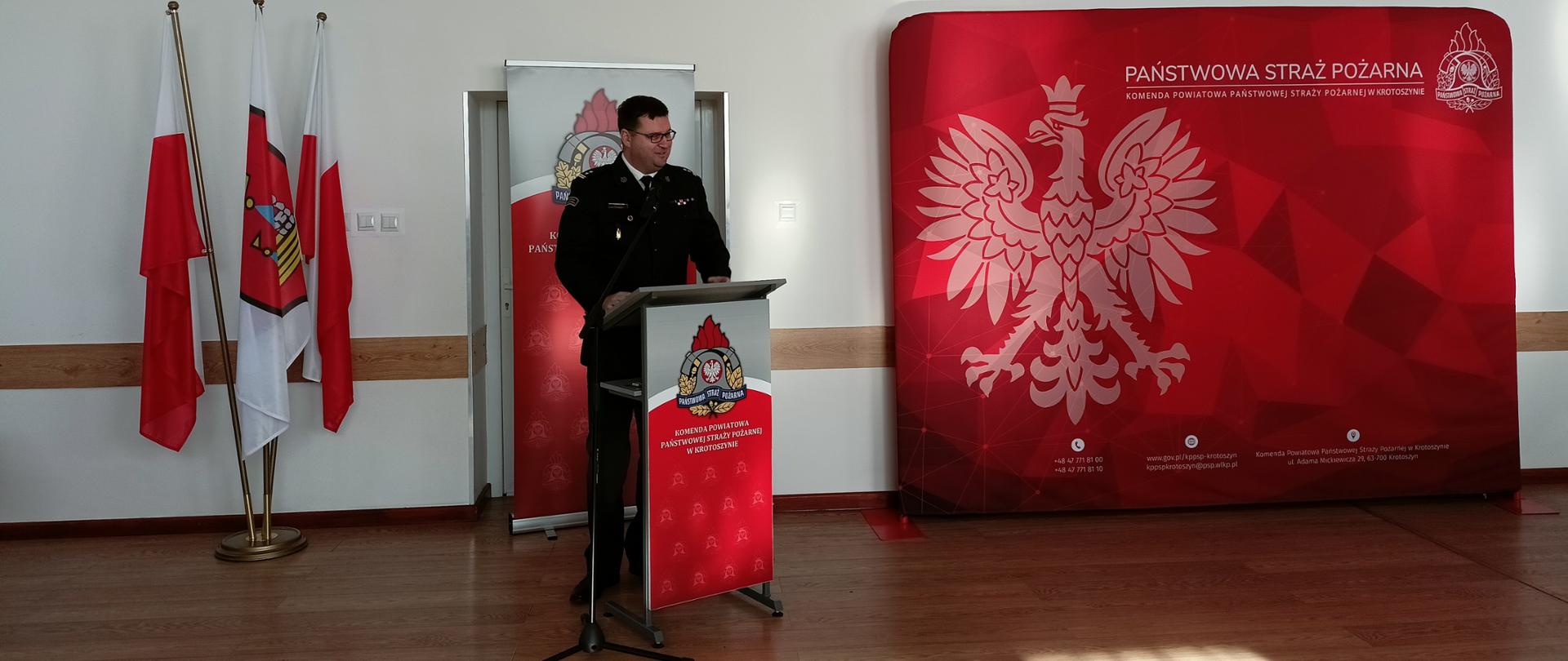 w dużej sali strażak stoi przy mównicy, przemawia, w tle duża ścianka tekstylna, czerwona z orłem, oraz flaga biało czerwona