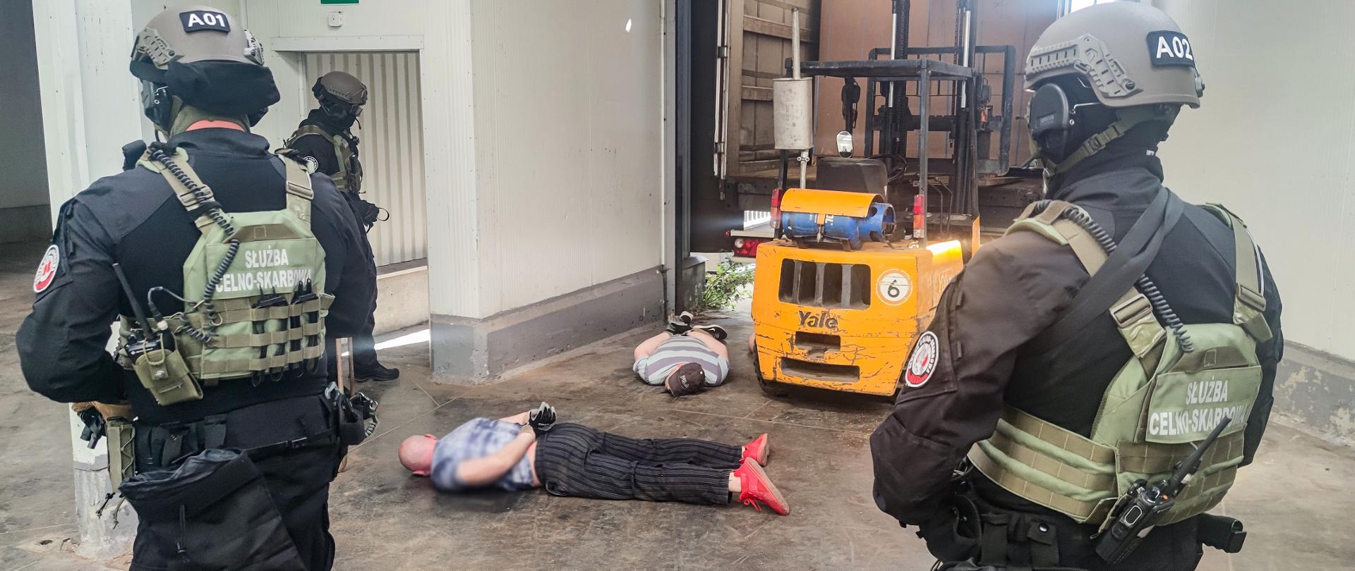 Zdjęcie przedstawia dwóch leżących na ziemi mężczyzn zatrzymanych przez funkcjonariuszy służby celno-skarbowej.