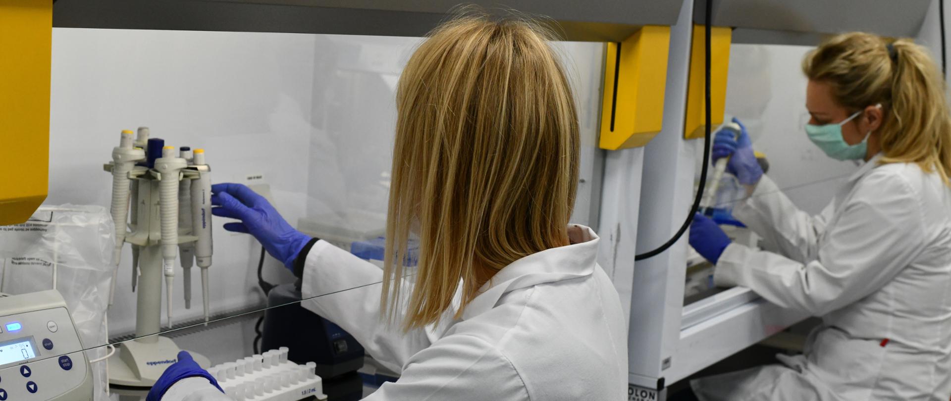 Zdjęcie przedstawia diagnostę laboratoryjnego podczas pracy. Na zdjęciu widoczne są 2 pracownice laboratorium podczas wykonywania pracy. Ubrane w białe fartuchy. 