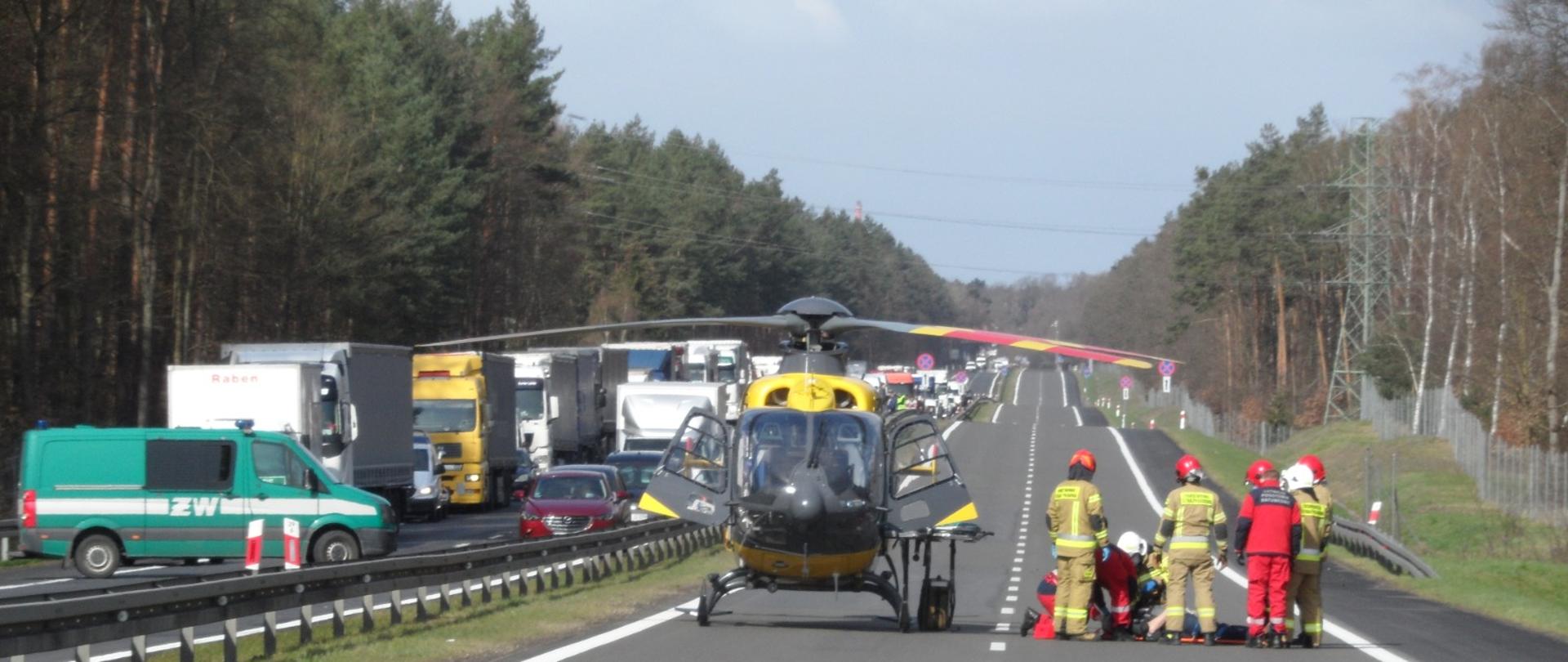 W centrum na pasie ruchu śmigłowiec LPR po prawo ratownicy LPR wraz ze strażakami po lewo zablokowany ruch przez pojazd Żandarmerii Wojskowej.