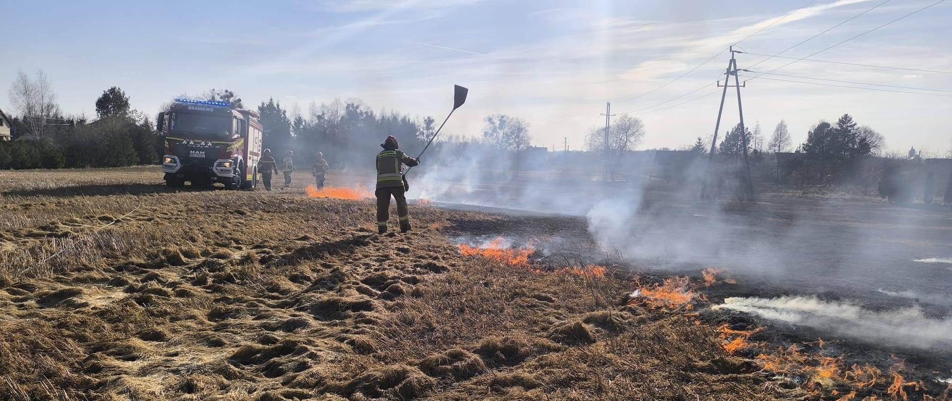 Strażacy gaszą pożar suchych traw. Widać połoninie. Z lewej strony u góry stoi samochód strażacki. Słonecznie.