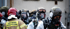 Policjanci w specjalistycznych strojach chemicznych ustawieni są jeden za drugim obok strażak w ubraniu specjalnym oraz hełmie sprawdza ich ubiór.