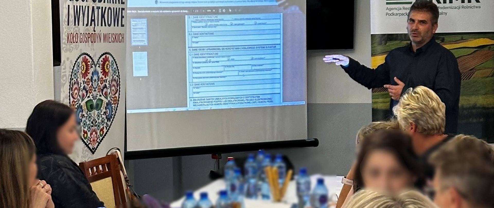 Ekspert stoi przy ekranie, na którym wyświetlana jest prezentacja. Przed nim siedzą przy stole uczestniczy szkolenia.