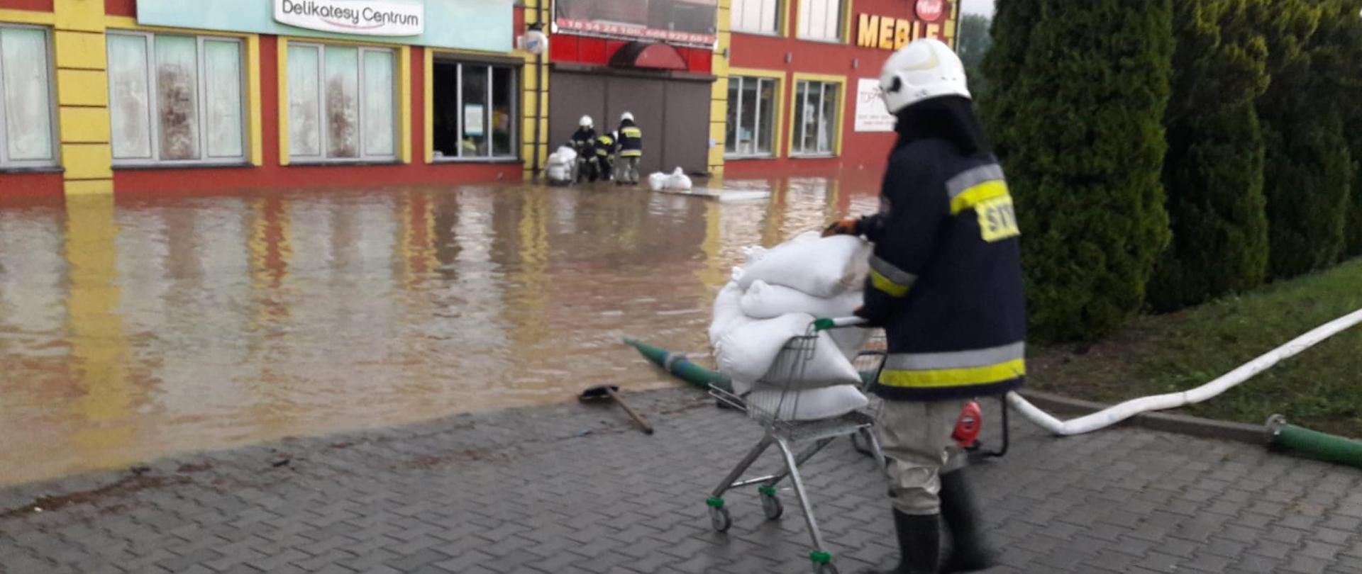 Strażak na wózku wiezie worki z piaskiem. W tle zalany plac a w wodzie strażacy prowadzący działania.