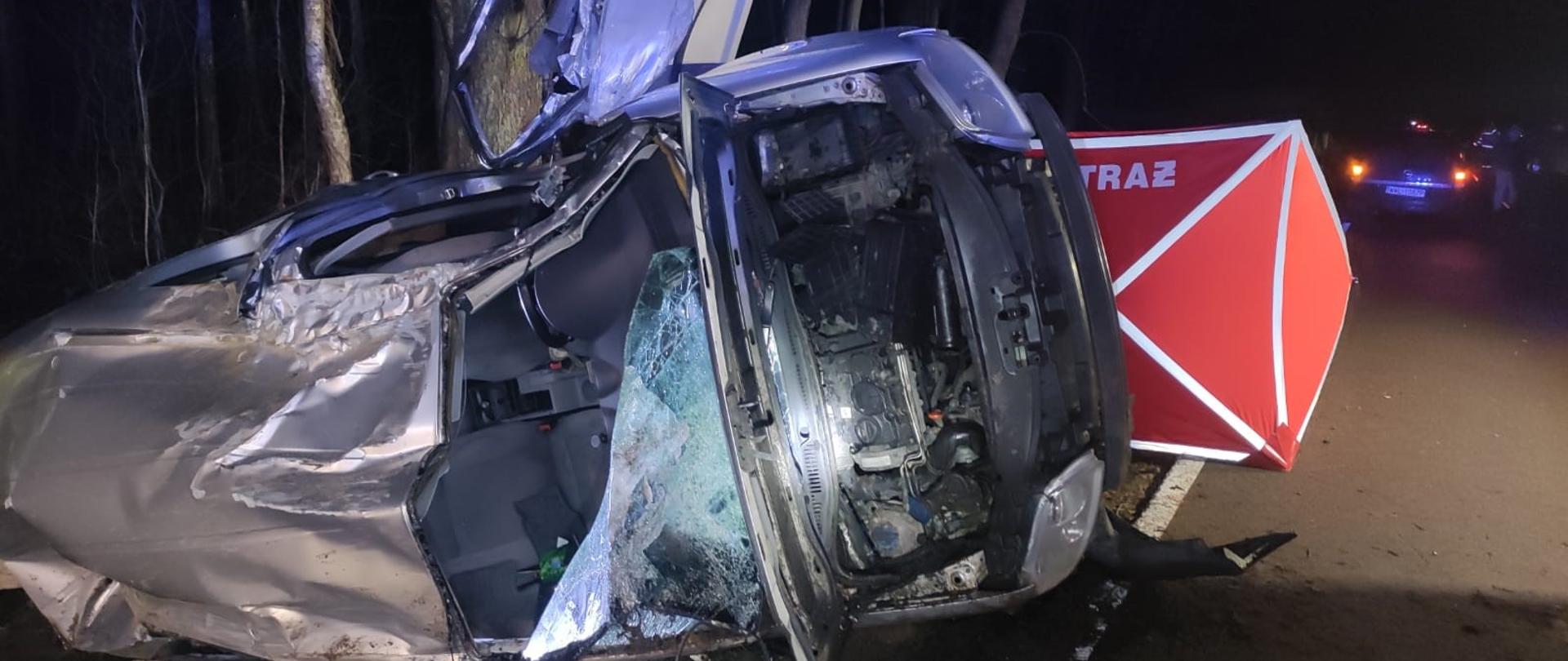 Na zdjęciu widoczny jest wrak samochodu osobowego leżący na prawym boku. Za pojazdem stoi czerwony strażacki parawan. W tle widać inny pojazd. Pora nocna.