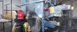 Na zdjęciu strażacy w piaskowych ubraniach i aparatach ochrony dróg oddechowych. Strażacy przy pomocy bosaka podręcznego wybierają spaloną celulozę. Jest duże zadymienie.