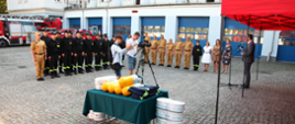 Strażacy Państwowej Straży Pożarnej ubrani w mundury kolory czarnego oraz piaskowego stoją wraz pracownikami cywilnymi na apelu obok ustawiony jest sprzęt pożarniczy pod namiotem stoi mężczyzna w garniturze przed nim stoją przedstawiciele prasy.