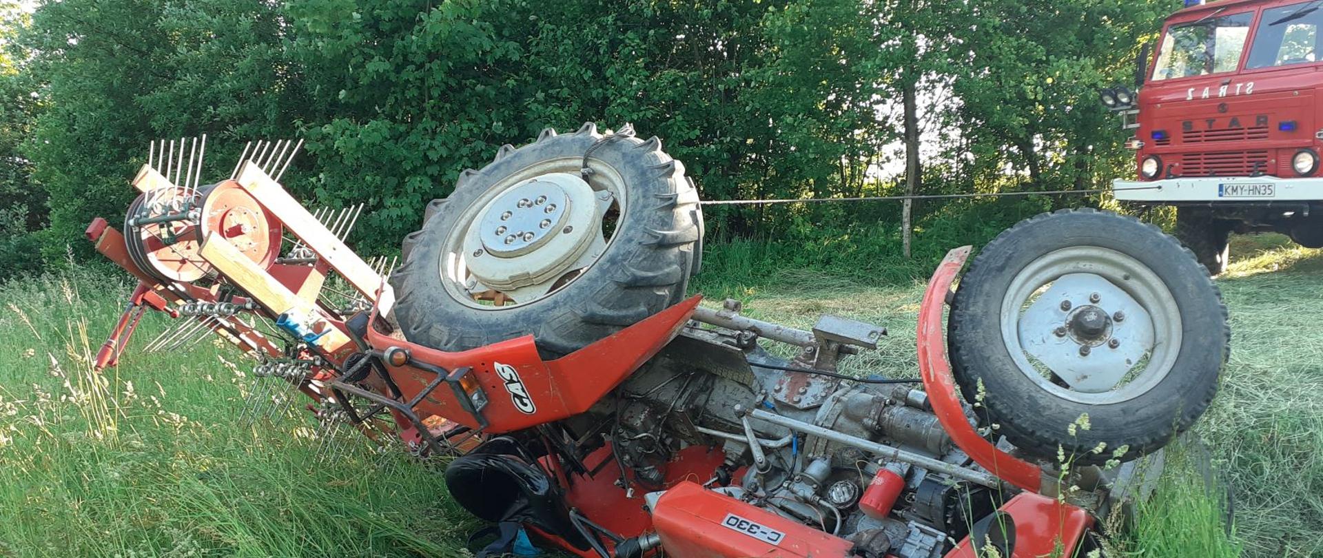 Na zdjęciu widoczny jest przewrócony ciągnik rolniczy podtrzymywany wciągarką samochodową. Znajduje się on na łące pokrytej trawą. Z prawej strony stoi samochód strażacki z napędem terenowym.