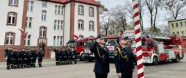 Poczet flagowy wciąga flagę Polski na masz pozostali strażacy ustawieni w dwóch rzędach oddają honory niedaleko stoją samochody gaśnicze.