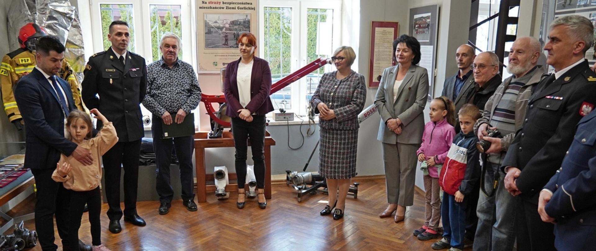 Na zdjęciu podczas wernisażu wystawy „Na STRAŻY bezpieczeństwa mieszkańców Ziemi Gorlickiej" widoczni w ubraniach galowych koloru granatowego strażacy, policjant i w ubraniach cywilnych zaproszeni na wernisaż goście 14 osób. W tle widoczne zdjęcia i eksponaty pożarnicze pompy, działko, węże pożarnicze, hełm, radiostacje, ubranie strażackie specjalne koloru pisakowego 