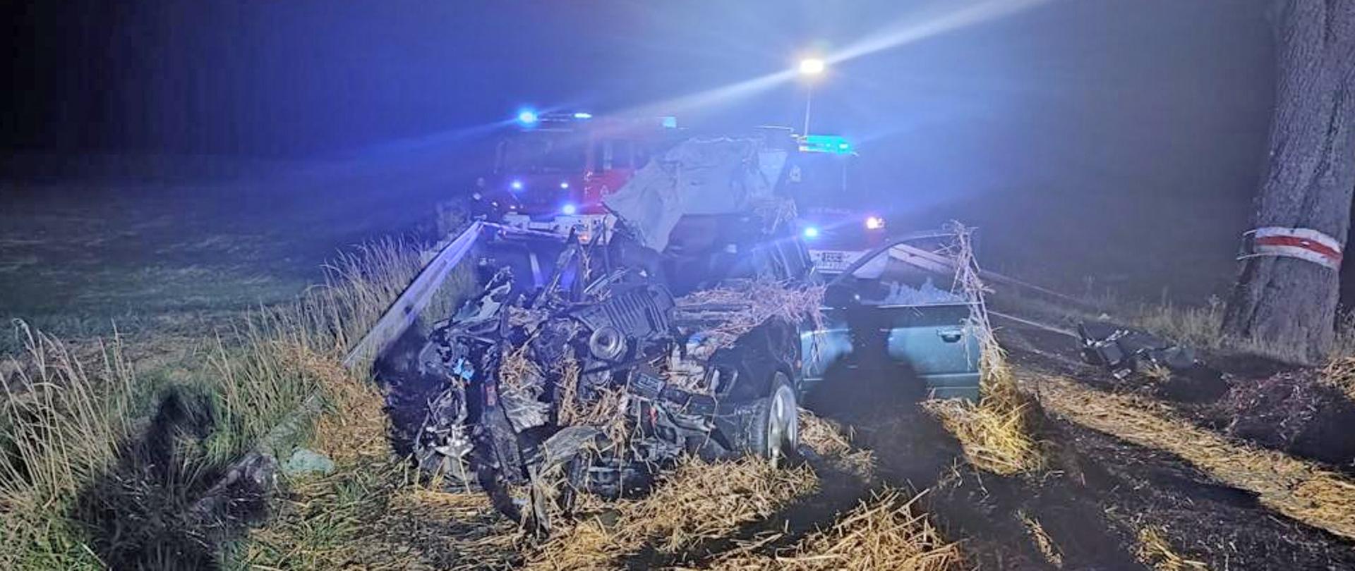 Na zdjęciu pora nocna, na poboczu drogi znajduje się uszkodzony samochód osobowy, w tle pojazdy straży pożarnej.