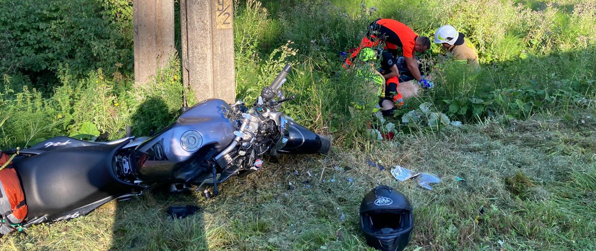 Na zdjęciu widać na pierwszym planie motocykl oraz kask motocyklowy. Motocykl leży przy słupie energetycznym, ma krzywą kierownicę, w pobliżu rozsypane są jego części oraz cały przód jest zniszczony. Około 2 metry dalej znajduje się 2 strażaków w ubraniach specjalnych, jeden to strażak psp a drugi osp, ratownik medyczny oraz poszkodowany motocyklista. Motocykliście udzielana jest pierwsza pomoc. Reszta na zdjęciu to zielone trawy i liście drzew. 