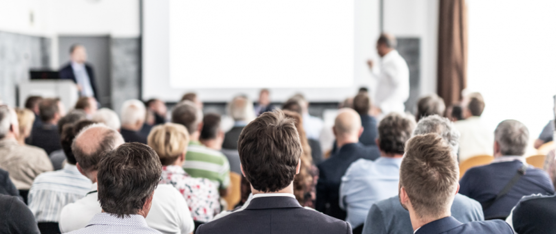 Audience seated in a conference room facing a presenter with a projection screen, with another person near a podium in the background.
