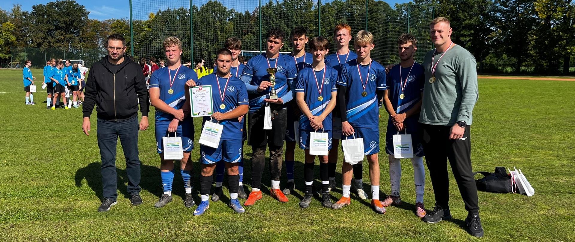 Drużyna w niebieskich strojach z medalami, pucharem, dyplomem i torbami upominkowymi stoi na boisku w słoneczny dzień. Po obu stronach stoją dwaj dorośli mężczyźni - przedstawiciel organizatora i opiekun grupy chłopców.