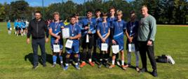 Drużyna w niebieskich strojach z medalami, pucharem, dyplomem i torbami upominkowymi stoi na boisku w słoneczny dzień. Po obu stronach stoją - przedstawiciel organizatora i opiekun drużyny.