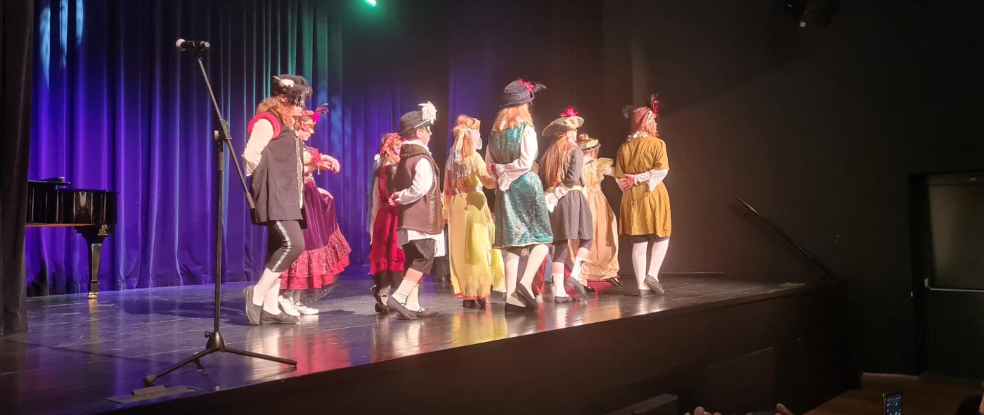 W pomieszczeniu - sala widowiskowa Ośrodka Kultury w Oławie. Na scenie, na tle czarnych kotar uczniowie Szkoły Muzycznej I st. w Oławie tańczą w strojach stylizowanych na stroje z epoki. 