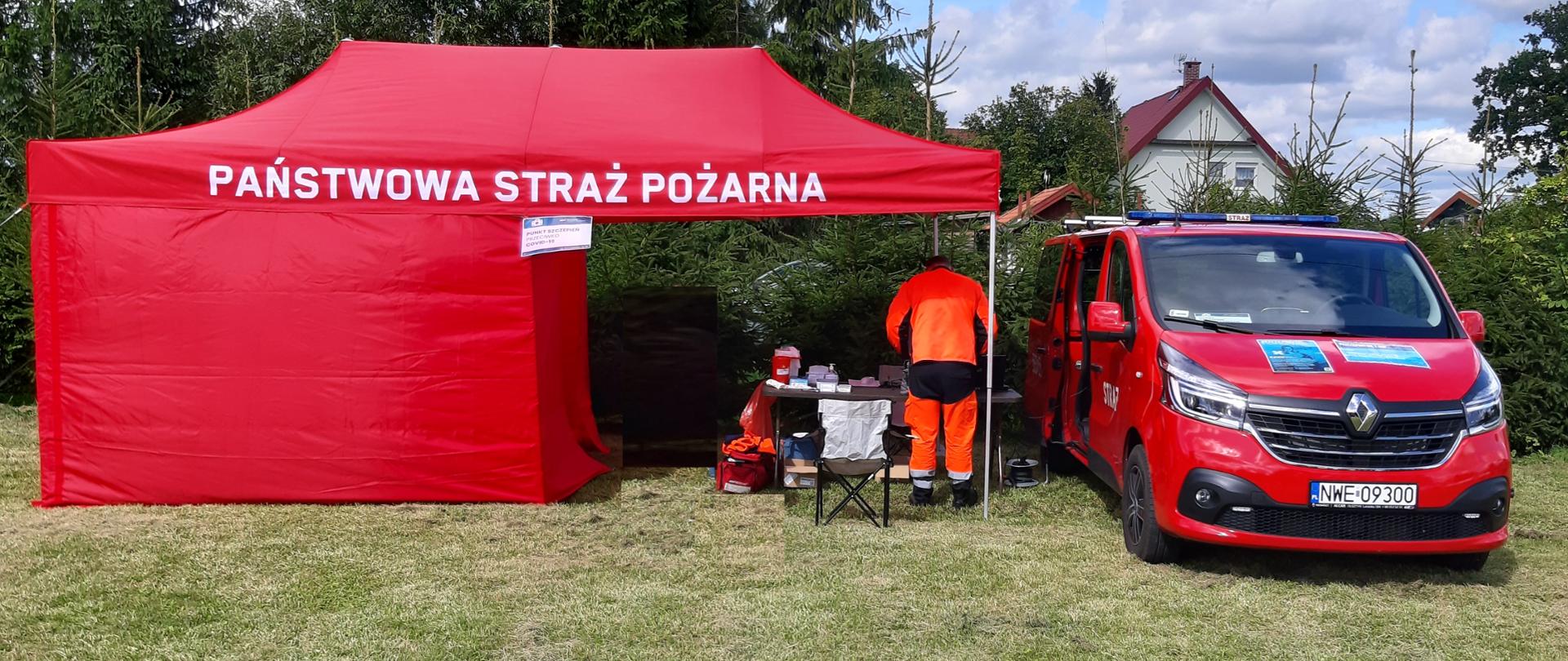 Na podwórku stoi namiot strażacki, w namiocie stoi ratownik medyczny, który przygotowuję stanowisko do szczepień. Obok niego stoi samochód strażacki. 