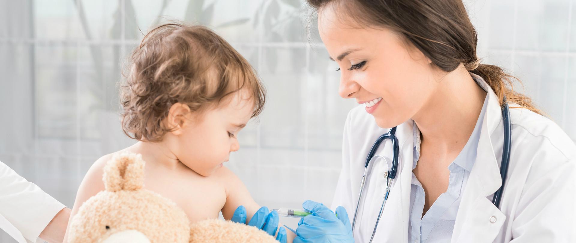 Uśmiechnięta młoda lekarka zaszczepia małe dziecko trzymające misia.