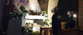 Zdjęcie przedstawia wnętrze kościoła, w którym przygotowano Grób Pański – tradycyjną dekorację upamiętniającą złożenie Jezusa do grobu.
Oto szczegółowy opis elementów widocznych na fotografii:
Centrum kompozycji: W centralnej części znajduje się ołtarz udekorowany licznymi bukietami białych kwiatów (prawdopodobnie goździków i chryzantem) oraz żółtych tulipanów. Pod ołtarzem umieszczona jest figura leżącego Chrystusa owinięta w całun.
Tło: Nad ołtarzem góruje duży obraz lub plansza przedstawiająca czarny krzyż z przewieszoną przez ramiona białą materią. Tło obrazu ma odcienie fioletu, różu i błękitu, przypominające niebo o zmierzchu. Po prawej stronie ołtarza, na podwyższeniu, widoczna jest ozdobna, złota monstrancja z Najświętszym Sakramentem, okryta półprzezroczystym tiulem.
Warta przy grobie: Po prawej stronie zdjęcia stoją dwaj strażacy w pełnym umundurowaniu galowym z napisem „STRAŻ” na plecach. Jeden z nich stoi tyłem, blisko ołtarza, drugi nieco dalej w głębi. Obaj mają na głowach błyszczące, złote hełmy i trzymają halabardy, pełniąc uroczystą wartę.
Architektura i oświetlenie: Scena rozgrywa się w bocznej nawie lub przy ołtarzu bocznym, co sugeruje zdobiony łuk i fragmenty polichromii na ścianach. Wnętrze jest słabo oświetlone, panuje w nim podniosły, refleksyjny nastrój. Światło skupia się głównie na kwiatach i białych elementach dekoracji, co kontrastuje z ciemnymi sylwetkami strażaków.