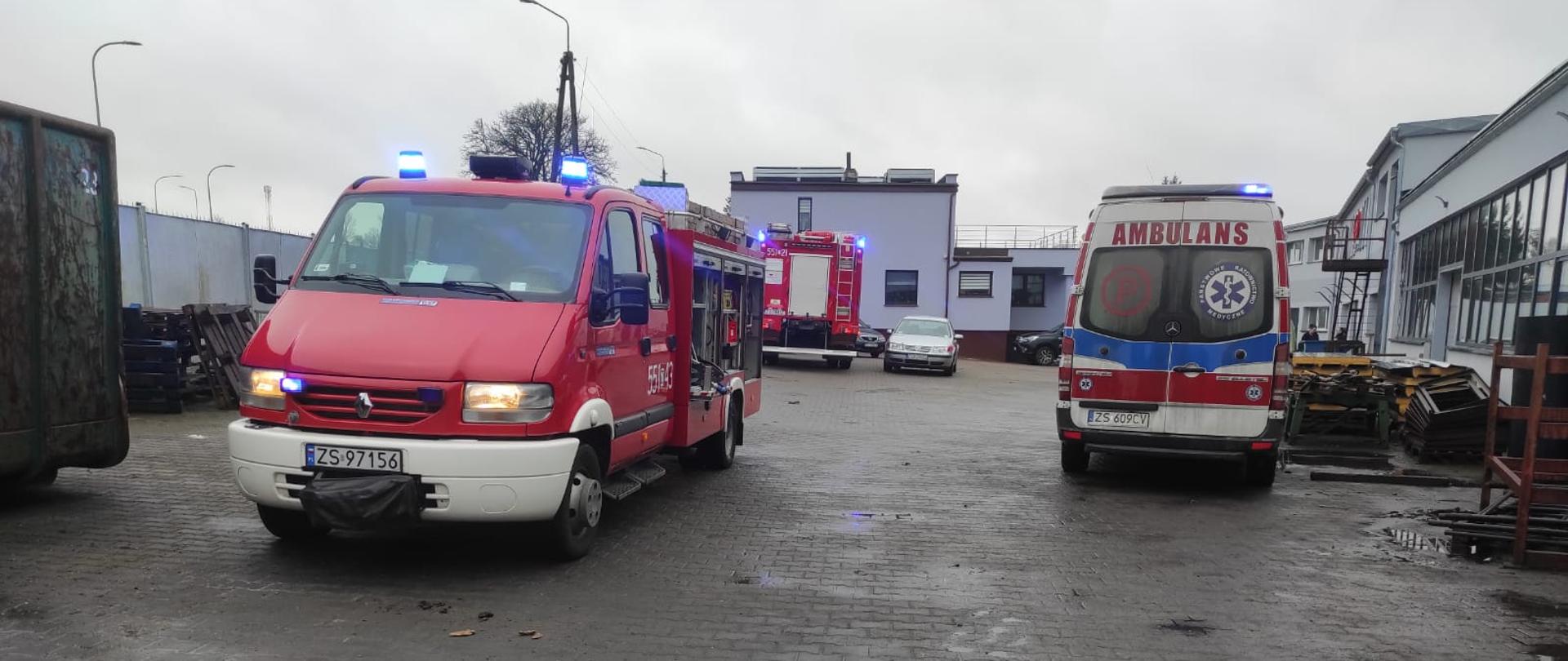Zdjęcie przedstawia wozy strażackie oraz zespół ratownictwa medycznego na placu przed zakładem produkcyjnym gdzie przygnieciony został mężczyzna