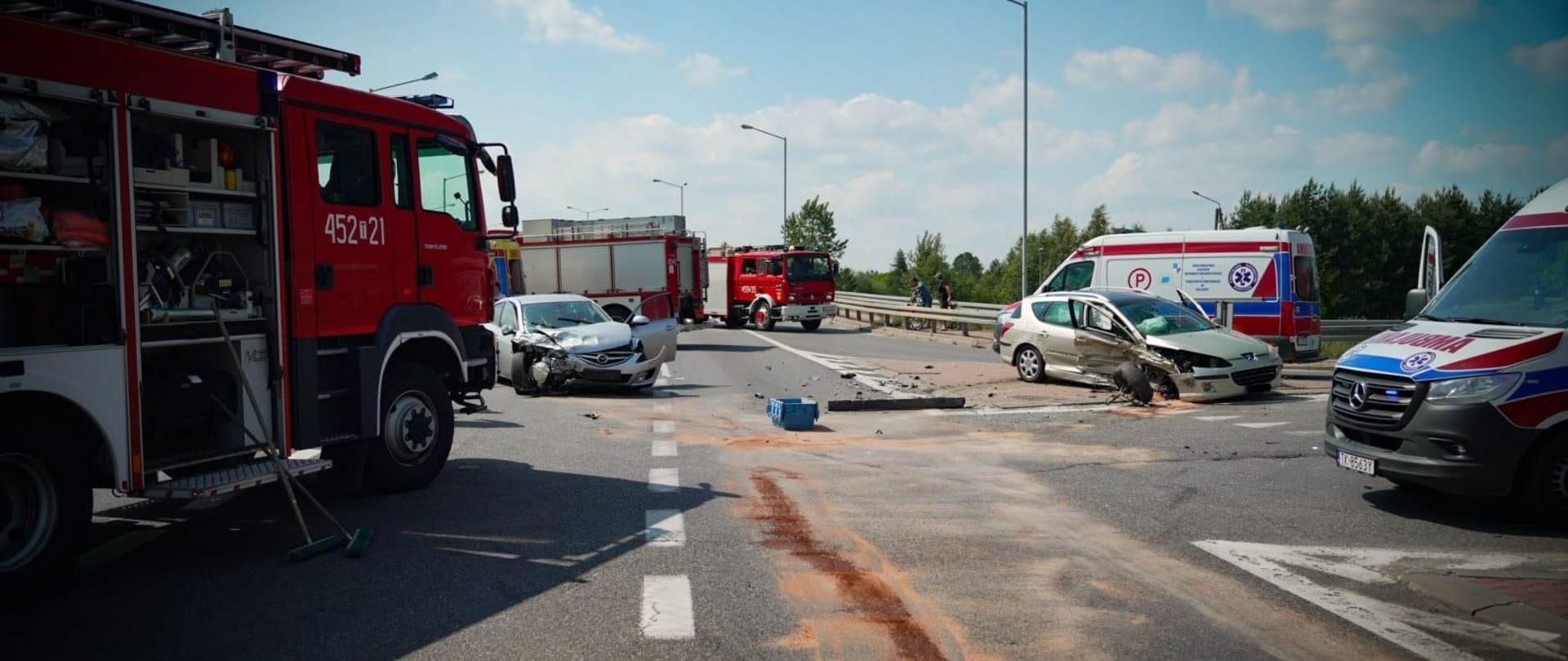 Na pierwszym planie po lewej widoczny jest samochód ratowniczo gaśniczy straży pożarnej, po prawej karetka pogotowia. Na drugim planie widoczne są dwa uszkodzone samochody osobowe. W oddali widoczne są dwa samochody ratowniczo gaśnicze oraz karetka pogotowia.