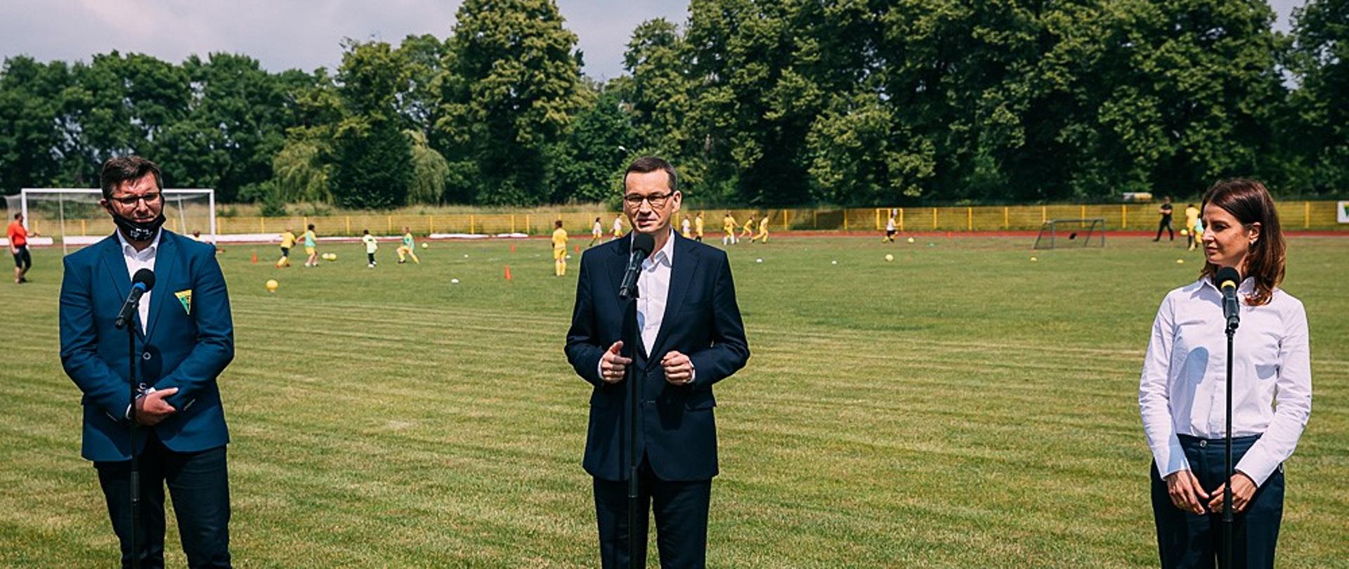 Minister Sportu Danuta Dmowska-Andrzejuk wraz z Premierem Mateuszem Morawieckim podczas konferencji na boisku piłkarskim.