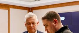 Na pierwszym planie widać dwie osoby. Po lewej ministra Tomasza Siemoniaka, natomiast po prawej Kyryło Budanowa, szefa Biura Prezydenta Ukrainy. Minister Tomasz Siemoniak wręcza upominek. Dodatkowo po prawej stronie widać niebieski ekran.