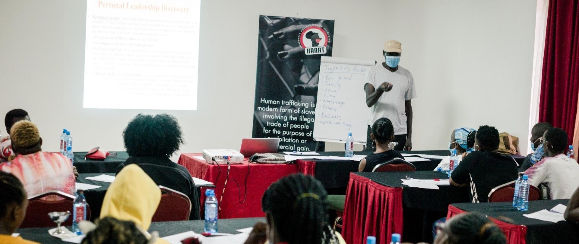 Ciemnoskórzy ludzie siedzą przy stołach. Na ścianie jest wyświetlana prezentacja. Jeden mężczyzna stoi.