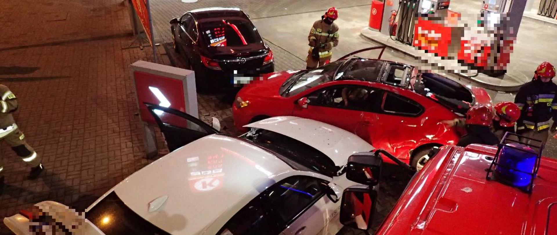dwa rozbite samochody osobowe na terenie stacji benzynowej, obok samochód straży pożarnej, wokół aut pracujący strażacy