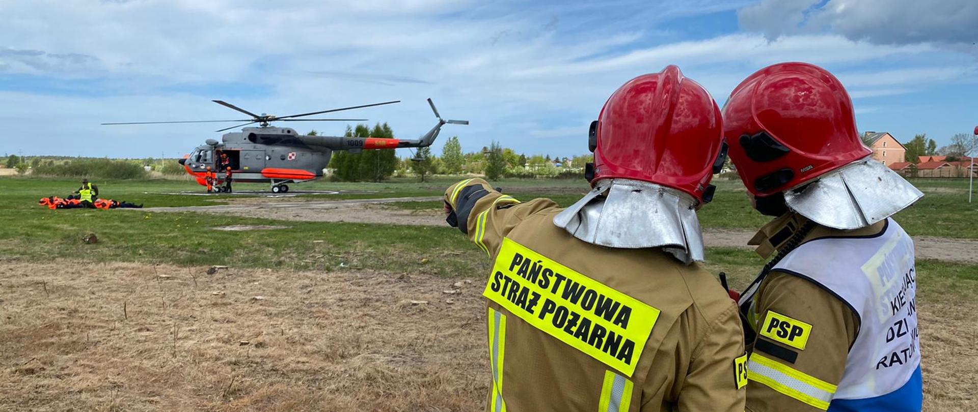 Zdjęcie przedstawia dwóch konsultujących się funkcjonariuszy Państwowej Straży Pożarnej zwróconych plecami do fotografującego na tle śmigłowca ratowniczego Mi14. Obok widoczne osoby poszkodowane przetransportowane przez śmigłowiec ratowniczy.