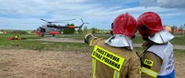 Zdjęcie przedstawia dwóch konsultujących się funkcjonariuszy Państwowej Straży Pożarnej zwróconych plecami do fotografującego na tle śmigłowca ratowniczego Mi14. Obok widoczne osoby poszkodowane przetransportowane przez śmigłowiec ratowniczy.