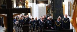 W ławach kościelnych stoją strażacy w mundurach galowych - widok na kilkadziesiąt osób. Wśród nich są też w dali cywile. Na pierwszej ławce leżą cztery czapki oficerskie. Od lewej, z przodu duży krzyż z Chrystusem, zwrócony do wiernych.