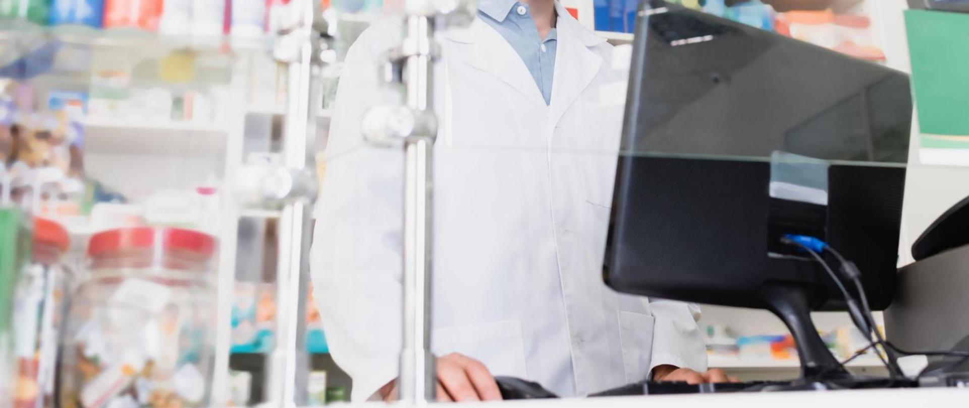 Na zdjęciu widać farmaceutę stojącego za ladą. W tle widać leki.
