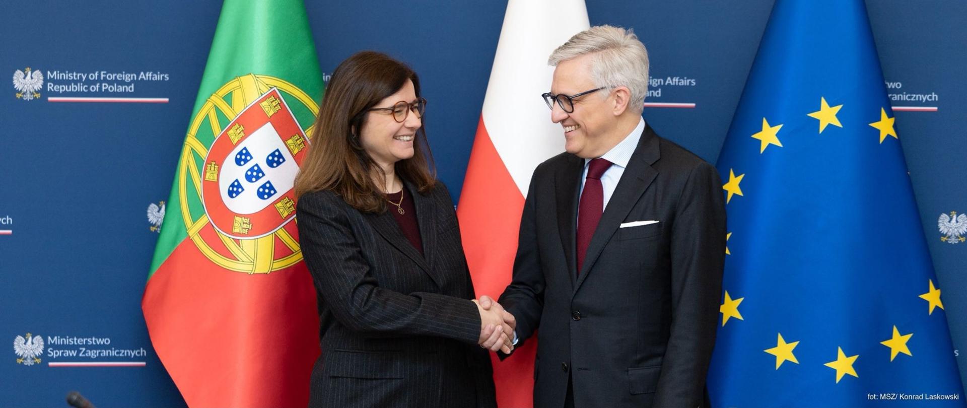 Inês Domingos, Secretária de Estado para os Assuntos Europeus no Ministério dos Negócios Estrangeiros de Portugal, e Embaixador Artur Harazim, Subsecretário de Estado no Ministério dos Negócios Estrangeiros da Polónia, trajados de forma formal, cumprimentam-se com um aperto de mão e trocam sorrisos. Ao fundo veem-se as bandeiras de Portugal, da Polónia e da União Europeia, bem como um painel azul com o logótipo do Ministério dos Negócios Estrangeiros da República da Polónia.