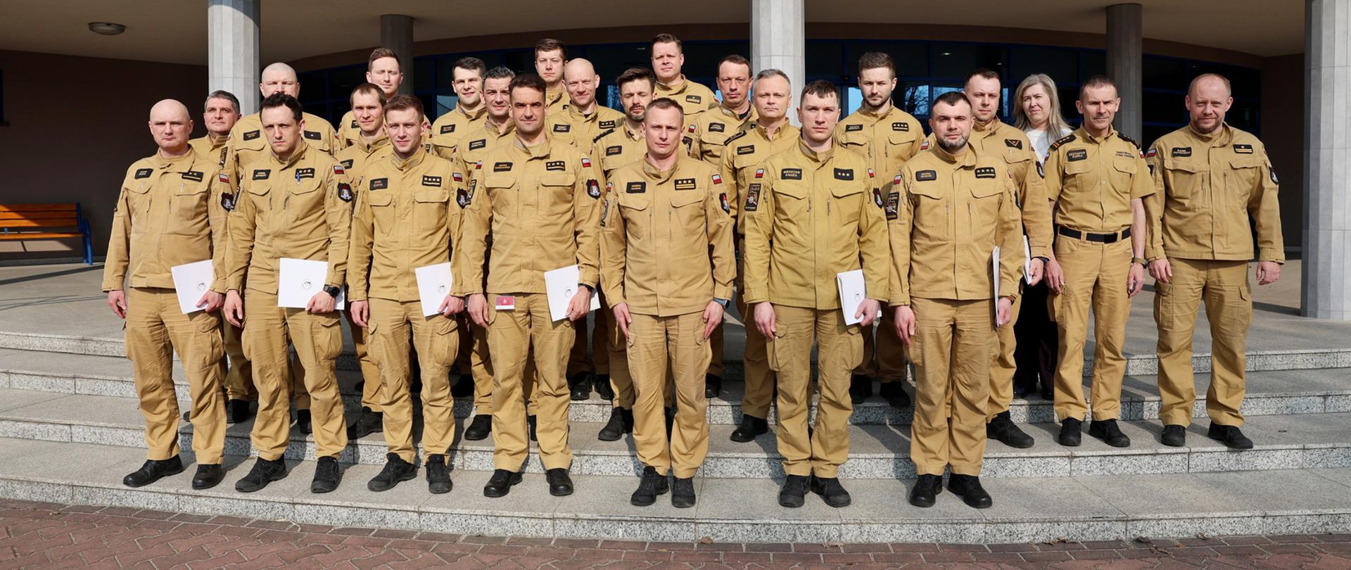 Pamiątkowe zdjęcie grupowe funkcjonariuszy PSP będących uczestnikami szkolenia z zakresu kierowania działaniami ratowniczymi na poziomie taktycznym (20 osób) w towarzystwie Zastępcy Komendanta Centralnej Szkoły PSP, kobiety oraz dwóch oficerów CS PSP