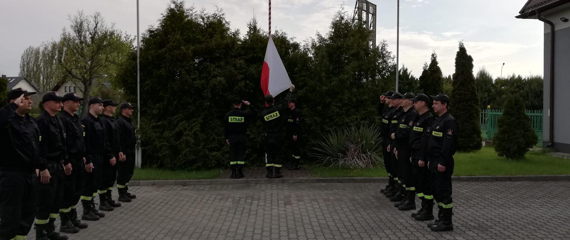 Na zdjęciu widać trzech strażaków, którzy zawieszają flagę Polski na maszcie. Po lewej i prawej stronie stoją strażacy w jednym rzędzie naprzeciwko siebie. W sumie jedenaście osób. Wszyscy ubrani są w czarne stroje z jasnożółtymi odblaskami z napisem straż. Na głowach strażacy mają założone czapki z daszkiem. Uroczysta zbiórka odbywa się na placu manewrowym przed budynkiem Komendy Powiatowej Państwowej Straży Pożarnej w Łęczycy. W tle widać krzewy. Jest widno, ale pochmurnie. 