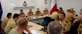 Zdjęcie przedstawia oficjalną odprawę kadry kierowniczej Państwowej Straży Pożarnej, odbywającą się w jasnej, nowoczesnej sali konferencyjnej. Około piętnastu strażaków siedzi wokół długich stołów ułożonych w kształt litery U. Wszyscy zebrani noszą jednakowe, piaskowe (beżowe) mundury służbowe z pagonami oznaczającymi stopnie i naszywkami z polską flagą na ramionach. Część z nich, siedząca na pierwszym planie, jest widoczna tyłem lub z profilu, skupiając się na dokumentach i dyskusji. Dwóch strażaków na pierwszym planie ma na szyi biało-czerwone, wzorzyste smycze.W głębi sali, na wprost obiektywu, siedzi czterech wyższych rangą oficerów, którzy aktywnie uczestniczą w naradzie – przed jednym z nich leży biały tablet, a pozostali przeglądają dokumenty lub uważnie słuchają.Na białej ścianie w tle, nad siedzącymi oficerami, wisi duża szklana tablica z logotypem formacji i napisem „PAŃSTWOWA STRAŻ POŻARNA KOMENDA WOJEWÓDZKA PAŃSTWOWA STRAŻ POŻARNEJ W KATOWICACH”. Po jej prawej stronie wisi godło Polski, a w narożniku pokoju stoi biało-czerwona flaga. Po lewej stronie kadru widoczny jest także fragment unijnej flagi. Atmosfera spotkania jest poważna i formalna